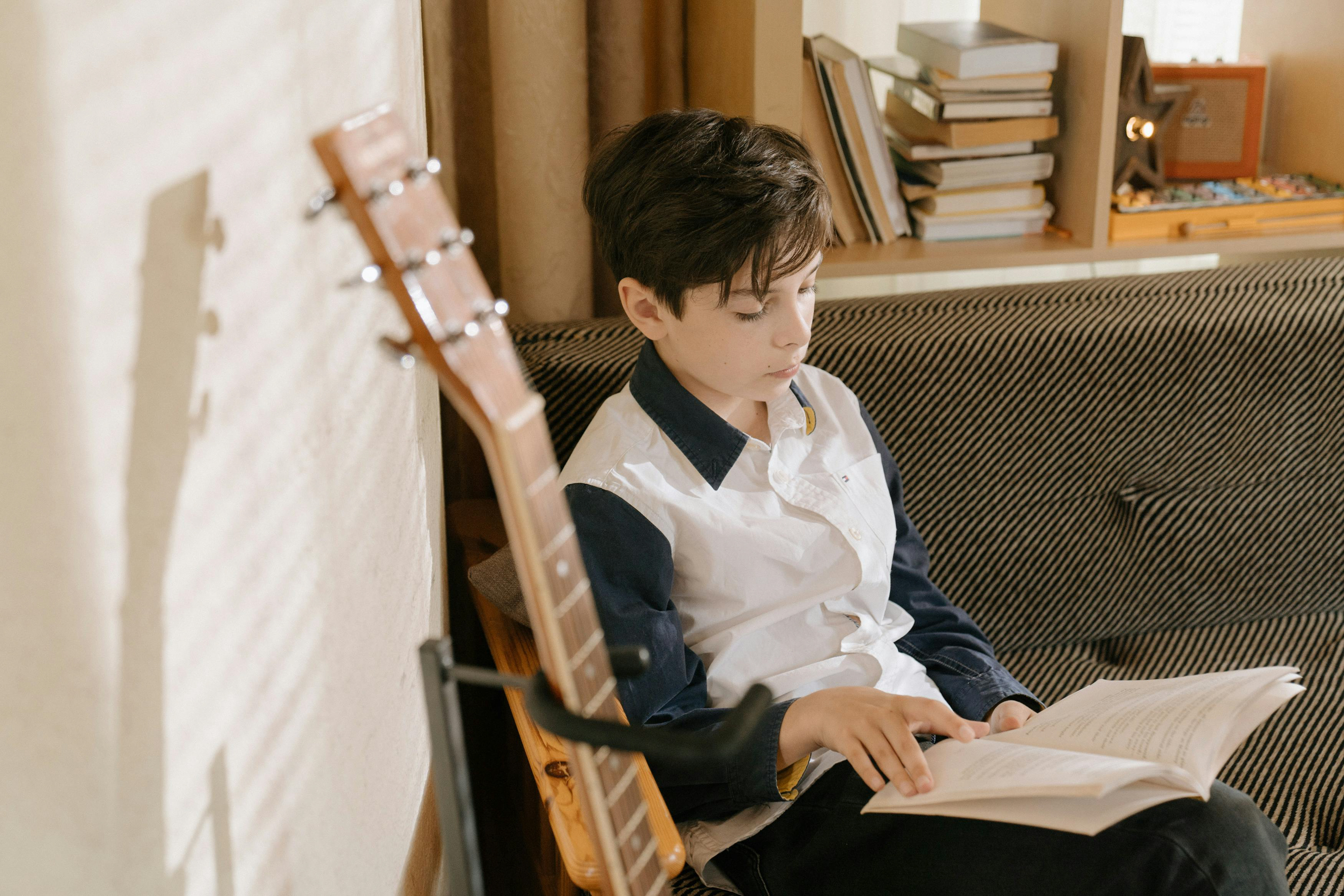 Boy sits on chair reading book next to guitar