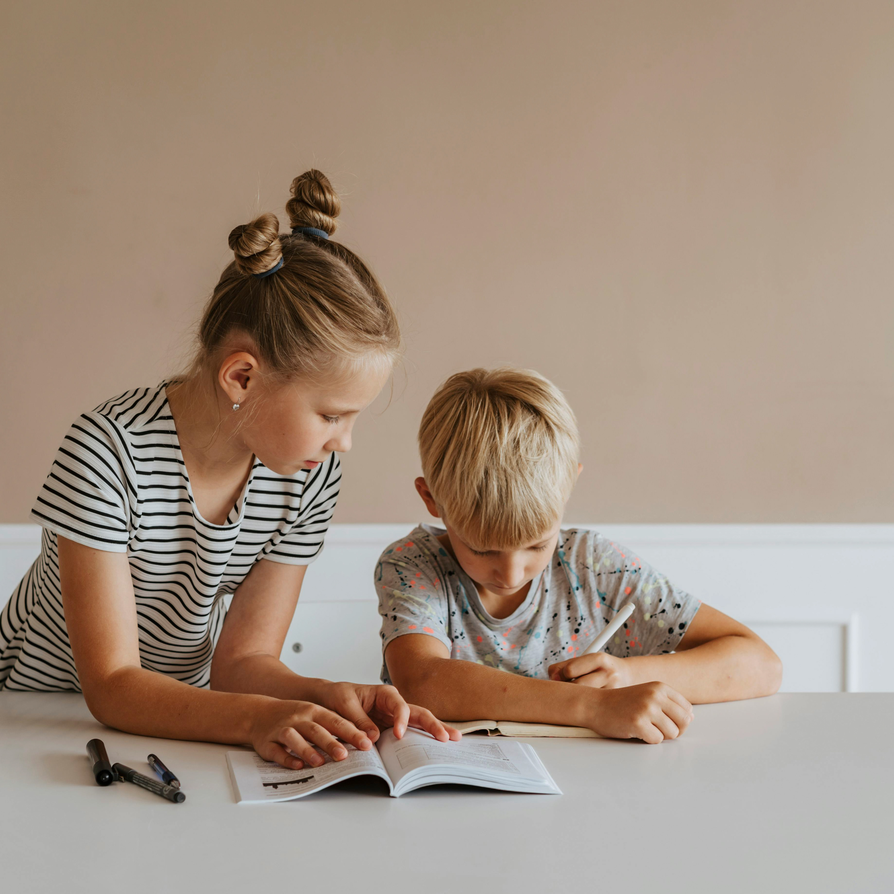 Two children sit at a table reading a book. One looks confused and is holding a pen